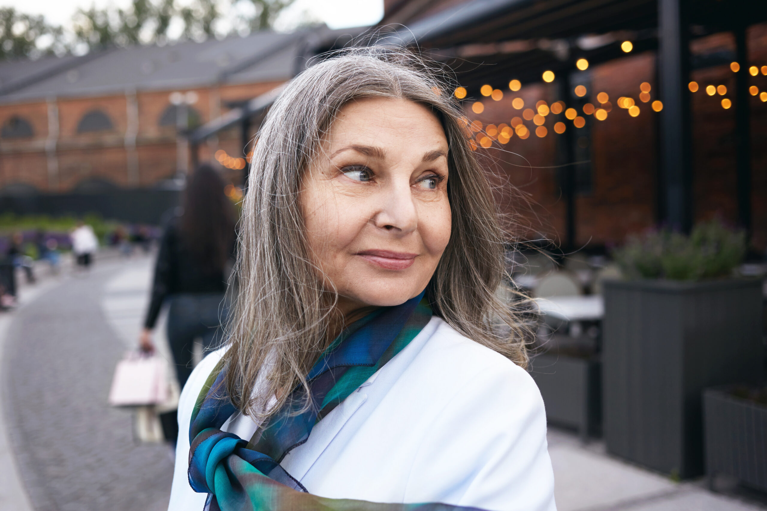 Urban lifestyle picture of beautiful happy mature woman wearing elegant clothes having curious excited facial expression while walking on street with blurred lights and people in background